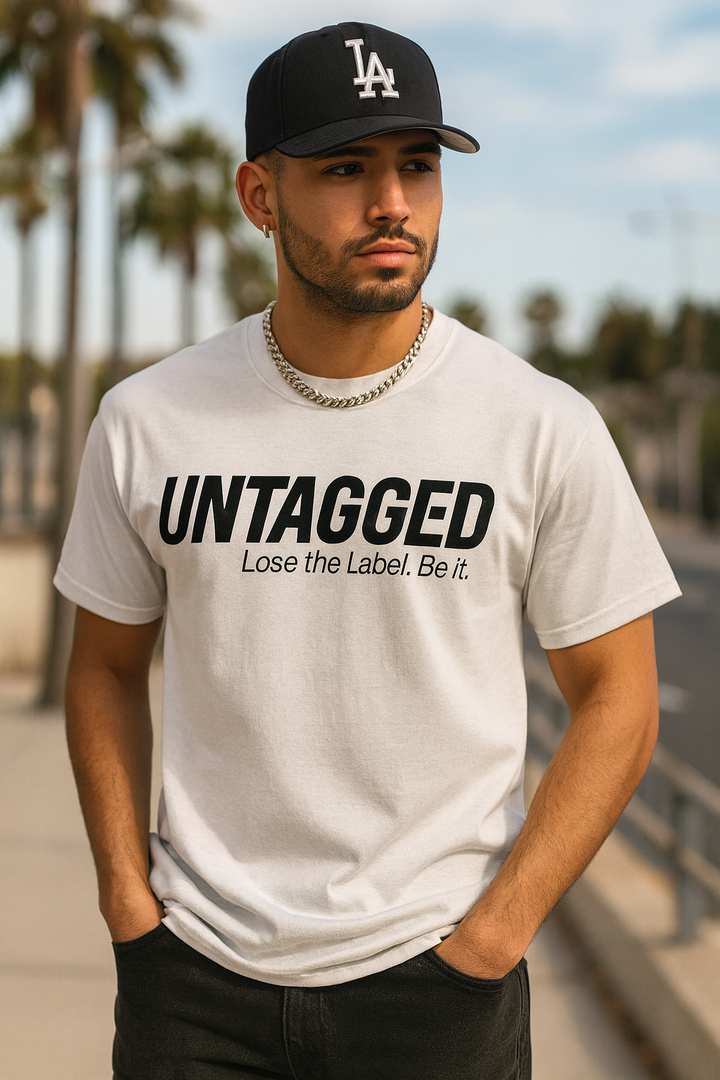 Man wearing a white t-shirt with 'UNTAGGED' text and a black cap outdoors.